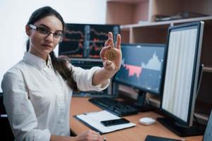 junger freiberufler in brillen. schöne frau, die bitcoin in den händen hält, während sie in einem modernen büro mit vielen monitoren mit grafiken sitzt foto