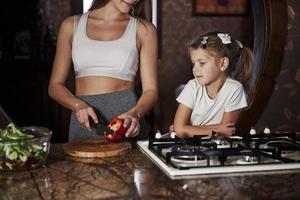 kind lernt kochen. hübsche junge frau, die in der modernen küche in der nähe des gasherds steht und der tochter beibringt, wie man essen zubereitet foto