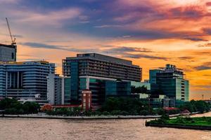 stadtbild des modernen gebäudes nahe dem fluss morgens bei sonnenaufgang. Bürogebäude der modernen Architektur in Thailand. Gebäude am Flussufer und Stadt mit orangefarbenem Himmel und Wolken foto