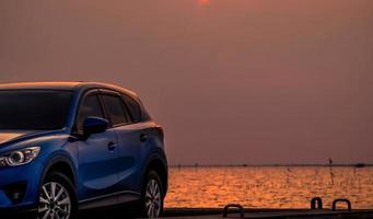 blaues kompaktes suv-auto mit sport und modernem design, das abends bei sonnenuntergang auf einer betonstraße am meer geparkt ist. hybrid- und elektroauto-technologiekonzept. Pkw-Stellplatz. Automobilindustrie. foto