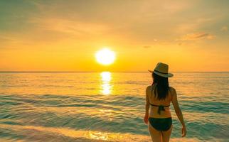 Silhouette erwachsene Frau, die am tropischen Meer mit wunderschönem Sonnenuntergangshimmel am Paradiesstrand spazieren geht. glückliches mädchen trägt bikini und strohhut entspannende sommerferien. Urlaubsreise. sommergefühl. Das Leben geht weiter. foto