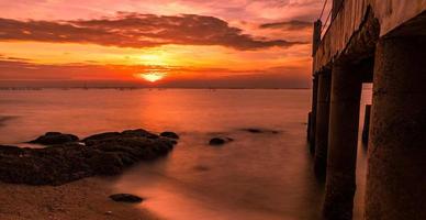 eine Langzeitbelichtungslandschaft mit wunderschönem Sonnenuntergang am Meeresstrand in der Nähe einer Betonbrücke mit orangefarbenem Himmel, Wolken und Stein foto