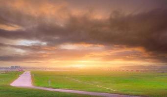 Landschaft der grünen Wiese am Flughafen. Unscharfe Start- und Landebahn mit Verkehrsflugzeugen, die starten oder landen, und orangefarbenem Sonnenlicht auf grauem, bewölktem Himmel. Luftfahrtgeschäft. Straße im Flughafen in der Nähe der Landebahn foto