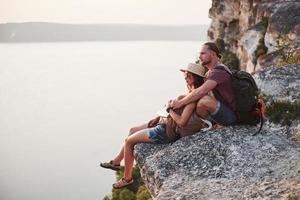 umarmendes Paar mit Rucksack, das oben auf dem Felsenberg sitzt und die Aussicht auf einen Fluss oder See genießt. Reisen entlang von Bergen und Küste, Freiheit und aktives Lifestyle-Konzept foto