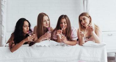 Frauencocktails trinken. Fröhliche Mädchen in der Nachtwäsche liegen auf dem Bett im weißen Raum und feiern foto