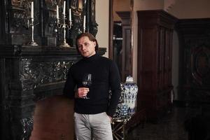 nachdenklicher Blick. junger schöner Mann in der Nähe des luxuriösen Kamins mit einem Glas Wein foto