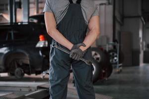 muskulöse Hände. Rückansicht des Arbeiters in Uniform, der mit einem Schraubenschlüssel in der Hand in der Werkstatt steht foto