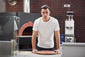 stylischer Typ. Bäcker im weißen Hemd mit Pizza, die zum Kochen in den Ofen gestellt werden kann foto