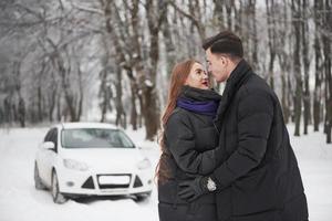 stylischer Mann. Wunderschönes junges Paar steht vor ihrem weißen Auto im verschneiten Wald foto