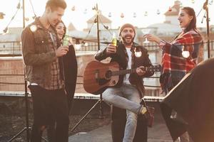 etwas Alkohol trinken. freunde haben spaß auf der dachparty mit dekorativen farbigen glühbirnen foto