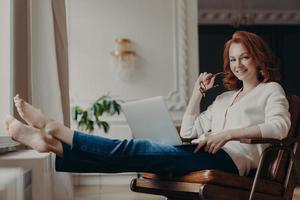 Fröhliche, erfolgreiche rothaarige Texterin arbeitet von zu Hause aus, hält den Laptop auf den Knien, streckt die Beine, denkt über eine Idee nach, um Werbeinhalte auf der eigenen Website zu erstellen, durchsucht Informationen im Internet foto