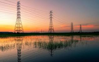 Hochspannungsmast und Elektrokabel mit Sonnenuntergangshimmel. Strommasten. Kraft- und Energiekonzept. Hochspannungsnetzturm mit Drahtkabel auf dem Reisfeld in der Nähe der Industriefabrik. foto