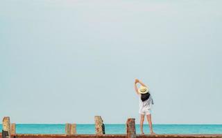 Rückansicht einer glücklichen jungen asiatischen Frau in lässiger Mode und Strohhut Entspannen Sie sich und genießen Sie den Urlaub am tropischen Paradiesstrand. Mädchen, das im Sommerurlaub am Holzsteg des Resorts steht. foto