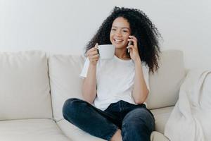 fröhliche afroamerikanerin hat kaffeepause im wohnzimmer, sitzt auf couch, ruft freund über modernes gerät an, hat glückliches lächeln, nachdenklichen ausdruck, bespricht etwas angenehmes. Technik, Lebensstil foto