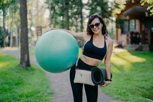 Fröhliche, sportliche junge Frau in Aktivkleidung hält einen großen Fitnessball, trägt eine Sonnenbrille, trägt Karemat, hat gute Laune, bereitet sich auf das Fitnesstraining im Freien vor, hat regelmäßige Sportübungen foto