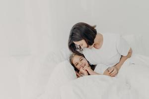 Fröhliche Mutter und Tochter haben gemeinsam Spaß im Bett, haben fröhliche Gesichtsausdrücke, genießen den Komfort weicher Bettwäsche, haben fröhliche Blicke, verbringen den Morgen im Schlafzimmer. Familien-, Erwachens- und Bettenkonzept foto