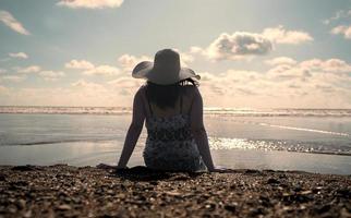 schöne junge hispanische frau, die allein am rand des strandes sitzt und einen hut und ein schwarz-weißes kleid während des sonnenuntergangs trägt foto