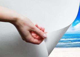 Hand, die eine Papierecke zieht, um aufzudecken, sonnigen Strand zu enthüllen foto