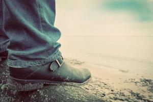Mann in Jeans und eleganten Schuhen, der auf einem umgestürzten Baum am wilden Strand steht und auf das Meer blickt. Jahrgang foto