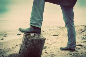 Mann in Jeans und eleganten Schuhen lehnt am Stammbaum am wilden Strand mit Blick aufs Meer. Jahrgang foto