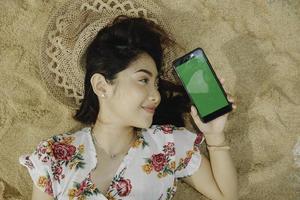 eine junge schöne asiatische frau, die auf dem sand liegt, während sie den telefonbildschirm zeigt foto