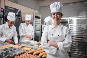 junge schöne asiatische köchin in weißer kochuniform blickt in die kamera, verschränkte arme und fröhliches lächeln mit professioneller berufstätigkeit in der lebensmittelbranche, kommerzielle gebäckküchenjobs in einer restaurantküche. foto