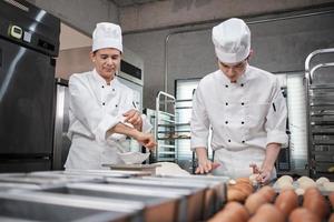 zwei professionelle asiatische männliche köche in weißen kochuniformen und schürzen kneten teig und eier, bereiten brot und frische backwaren zu und backen im ofen in der edelstahlküche des restaurants. foto