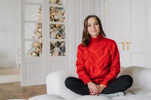 Foto einer angenehm aussehenden jungen Frau in rotem Pullover, Hose, sitzt mit gekreuzten Beinen auf einer bequemen Couch, genießt häusliche Atmosphäre, geschmückter Neujahrsbaum im Hintergrund. Home-Konzept