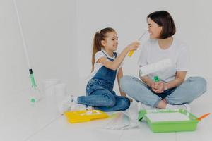 positives mädchen schmiert mütter nase mit farbe durch roller, viel spaß bei der hausrenovierung, sitzen zusammen auf dem boden im weißen raum, verwenden die notwendigen werkzeuge für die reparatur. Hausrenovierungskonzept foto