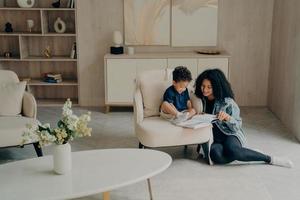 kleiner Mischlingsjunge, der mit seiner liebevollen Mutter liest, während er die Zeit zusammen im Wohnzimmer zu Hause genießt foto