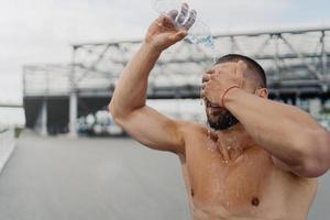 Der erschöpfte Sportler spritzt frisches kaltes Wasser über den Kopf, versucht sich nach hartem Training im Freien zu erfrischen, posiert ohne Hemd, hat starke Muskeln und eine perfekte Körperform. trink- und sportkonzept foto