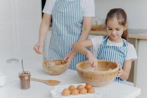 mutter gibt kleinem kind kochunterricht, steht nebeneinander, mischt zutaten in großen holzschüsseln, macht zusammen teig, verwendet eier, mehl und andere produkte. Familie, Kochen, Mutterschaft foto