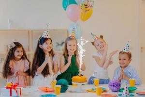 kinder- und urlaubskonzept. froh, dass fünf freunde freudig auf kuchen mit funkeln schauen, geburtstag feiern, partykegelhüte tragen und luftballons halten, fröhliche ausdrücke haben foto