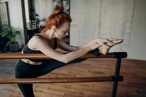 Schlanke rothaarige Frau, die sich im Klassenzimmer auf der Ballettstange dehnt foto