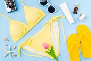 Draufsicht auf gelben Bikini und Cremeflasche, Sandalen, Sonnenbrille mit Strandzubehör auf blauem Hintergrund. Sommerzeit-Konzept. foto