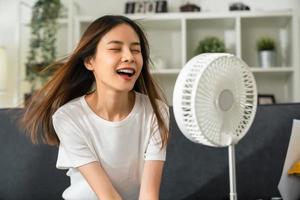 junge lächelnde asiatin, die auf der couch sitzt und den ventilator anschaltet, um sich abzukühlen, leidet unter zu heißem wetter im wohnzimmer. foto