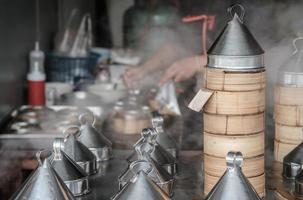 heißer, dampfender Dim-Sum-Bambuskorb, gestapelt auf rostfreiem Dampfgarer, mit Frau, die im Restauranthintergrund kocht. foto