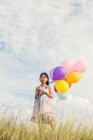 süßes kleines mädchen, das bunte luftballons auf der wiese gegen blauen himmel und wolken hält und die hände ausbreitet. foto