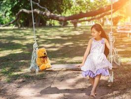 süßes kleines mädchen sitzt schaukeln, teddybär sitzt mit ihr, kleines mädchen zeigte auf den baum. foto