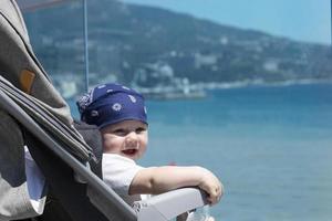 Nahaufnahme eines niedlichen sieben Monate alten Babys in einem Kopftuch, das vor dem Hintergrund der Stadtböschung und des Meeres lächelt. foto