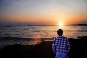 Porträt eines gutaussehenden Mannes, der auf einem Stein an der Seite des Meeres steht und mit einem Sonnenaufgangs-Abenteuerreisekonzept weit über das Meer blickt. foto