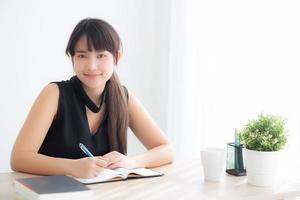 schöne junge asiatische frau, die lächelnd sitzend studiert und lernt, notizbuch und tagebuch im wohnzimmer zu hause zu schreiben, mädchenhausaufgaben, geschäftsfrau, die am tisch arbeitet, bildungskonzept. foto