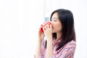 schöne junge asiatische frau des porträts mit trinken einer tasse kaffee stehendem vorhangfensterhintergrund im schlafzimmer, mädchen entspannen morgens zu hause, lebensstilkonzept. foto