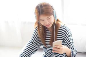 schöne asiatische junge frau genießt es, musik mit kopfhörern zu hören und ein intelligentes handy zu halten, während sie im schlafzimmer sitzt, entspannen sie sich mädchen mit kopfhörer, freizeit- und technologiekonzept. foto