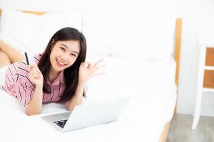schön von Porträt junge asiatische Frau liegend Benutzer Kreditkarte mit Laptop, zufriedenes Mädchen, das online einkauft und Zahlung mit Notebook-Computer auf dem Bett im Schlafzimmer, Lifestyle-Konzept. foto