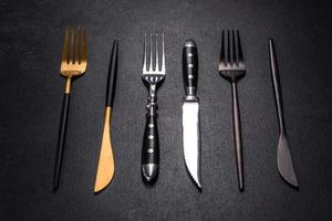 Mehrere Messer und Gabeln in Schwarzgold und Silber auf dunklem Betongrund