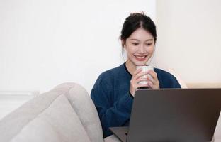 Portrait schöne junge Asiatin, die online am Laptop arbeitet, das Internet nutzt und die Tasse hält. zu hause auf dem sofa sitzen, freier platz foto