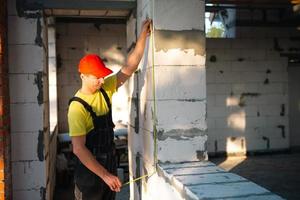 bauarbeiter auf der baustelle misst die länge der fensteröffnung und der mauer mit einem maßband. Häuschen bestehen aus porösen Betonblöcken, Arbeitskleidung - Overall und Baseballkappe foto