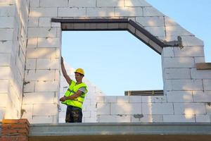 Bauarbeiter auf der Baustelle misst die Länge der Fensteröffnung und der Wand mit Maßband. Cottage bestehen aus Porenbetonblöcken, Schutzkleidung - Helm und Weste foto