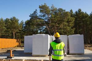 der projektarchitekt steht mit dem rücken auf der baustelle eines hauses, dessen fundament gelegt und die steine geliefert wurden. Bauarbeiter mit gelbem Schutzhelm und Signalweste. Attrappe, Lehrmodell, Simulation foto